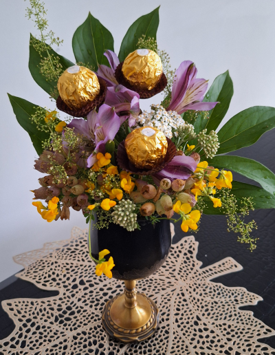 A vibrant mixed bouquet featuring orange lilies, red roses, and bright sunflowers, arranged with green foliage in a clear glass vase.