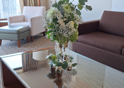 A white and green floral arrangement in a gold vase with crystal accents, displayed on a glass coffee table in a modern hotel suite with soft, neutral decor.