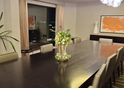 A luxurious meeting room with a long dark wood table, featuring an elegant floral arrangement of white and green flowers in the center, complemented by modern decor and a large window with a city view.