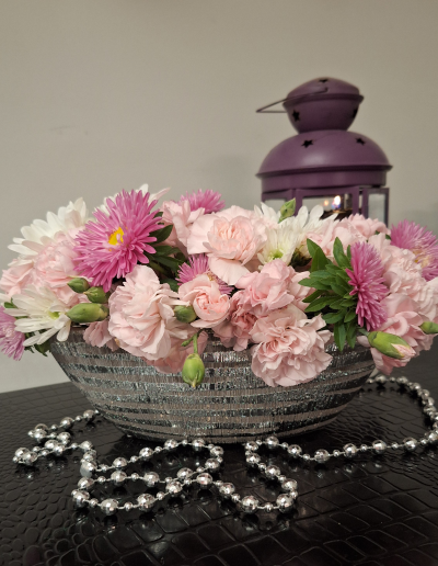 Flower arrangement in a silver-striped bowl with pink and purple blooms, surrounded by a silver beaded necklace.