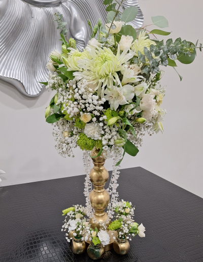 A stunning flower arrangement featuring white lilies, chrysanthemums, baby's breath, and green accents, elegantly displayed on a tall gold stand with cascading crystal beads.