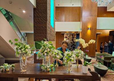 A florist arranging white and green floral centerpieces on a wooden table in a luxurious hotel lobby, featuring gold accents and crystal details.