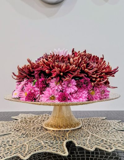 Layered floral arrangement featuring pink and deep red chrysanthemums displayed on an intricate gold stand, sitting on a textured leaf-patterned mat