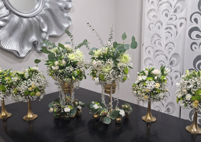 A collection of elegant white and green floral arrangements with roses, chrysanthemums, and eucalyptus displayed in gold vases with crystal accents, set against a modern background.