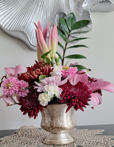 A bright floral arrangement featuring pink lilies, burgundy chrysanthemums, white carnations, and pink daisies, set in a silver vase with a reflective, aged texture.