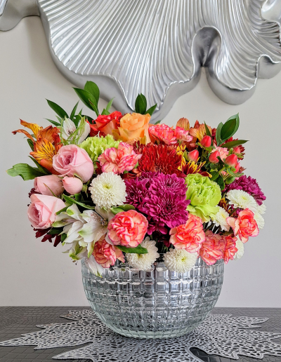 A large floral arrangement in a clear patterned glass bowl, featuring a mix of pink, orange, and green flowers.