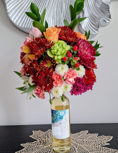 A colorful flower arrangement in a wine bottle, featuring roses, carnations, chrysanthemums, and greenery.