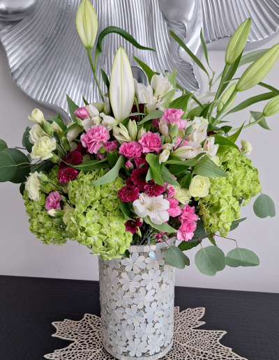 A close-up of a floral arrangement featuring green hydrangeas, white lilies, pink and purple carnations, and greenery in a decorative mosaic vase.