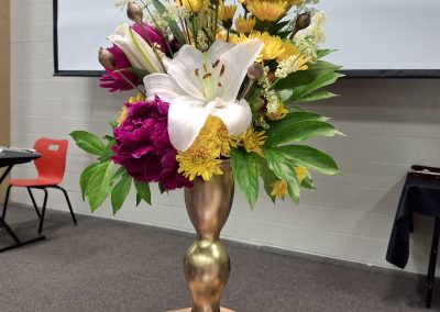 Close-up of a vibrant floral arrangement featuring white lilies, pink peonies, and yellow chrysanthemums in a gold vase, created by Pria Flowers for a Grade 6 graduation ceremony.