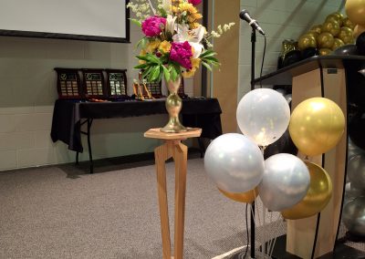 Floral arrangement by Pria Flowers displayed on a wooden pedestal beside a podium and microphone, decorated with balloons for St. Kateri Catholic School's Grade 6 graduation.