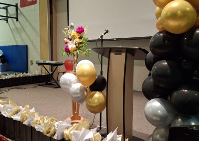 Wide view of the graduation stage at St. Kateri Catholic Elementary School featuring Pria Flowers’ floral arrangement, podium, gold and white balloons, and graduation gift bags.