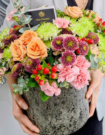 A person holding a modern flower arrangement with peach roses, pink carnations, chrysanthemums, red berries, and eucalyptus in a textured gray pot.
