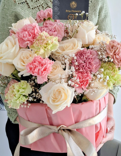 A person holding a pink flower box arrangement with white roses, pink carnations, baby’s breath, and soft greenery, tied with a ribbon.